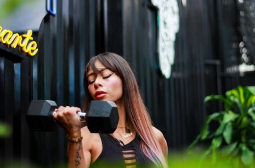 Fit woman with long hair and tattoos lifting a dumbbell with intense focus outdoors.